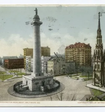 The Washington Monument rises from a public square, with surrounding city buildings and a gothic-style church steeple nearby.