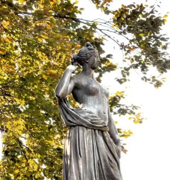 Weathered Diana Fountain statue seen from below against dense green foliage.