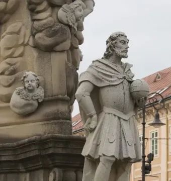 Weathered stone statue beside the Marian Column, with an ornate base decorated with cherub heads.