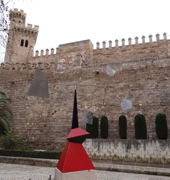 Nancy abstract sculpture with a red and black base in front of a stone fortress wall and trimmed hedges.