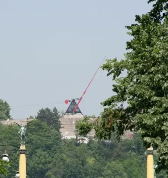 The Prague Metronome, a large red kinetic sculpture, stands on a stone pedestal above a tree-covered hill.