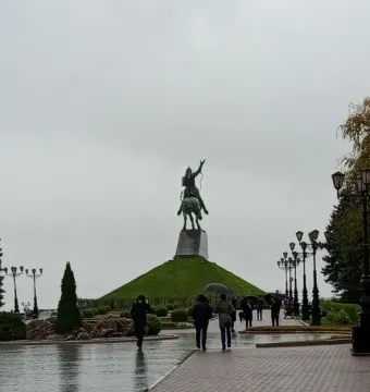 The Salavat Yulaev Monument stands on a grassy mound above a wet walkway, with visitors holding umbrellas on a rainy day.
