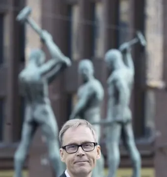 A smiling man in glasses and a suit stands in front of the Three Smiths sculpture.