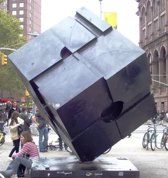 The Alamo, a dark cube sculpture, stands balanced on one corner in an urban plaza with buildings and traffic lights behind it.
