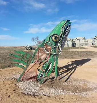 Carhenge-like roadside monument with cars arranged on gray supports behind a large green metal sculpture in the foreground.
