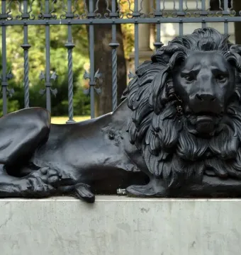 Guarding Lion sculpture on a concrete pedestal in front of an ornate fence and brick pillar.