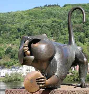 Heidelberg Bridge Monkey sculpture crouching on a stone ledge, holding a round disk with the river and wooded hill behind it.