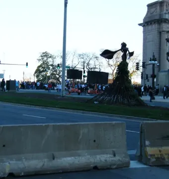 The Bowman statue stands on a stone pedestal beside a monumental archway, overlooking a street with barriers, traffic lights, and people.