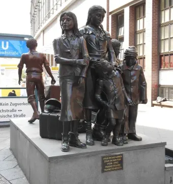 Trains to Life – Trains to Death bronze sculpture with human figures on a pedestal near a subway station entrance