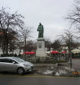 Vodnik Monument on a tall stone pedestal in a paved square with bare trees and buildings nearby.