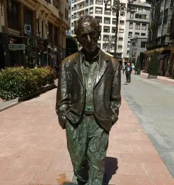Woody Allen memorial statue of a bespectacled man on a pedestrian walkway with city buildings behind it.