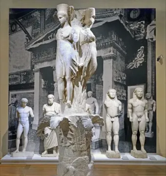 Dancers of Delphi sculpture with three draped female figures atop a textured column base.
