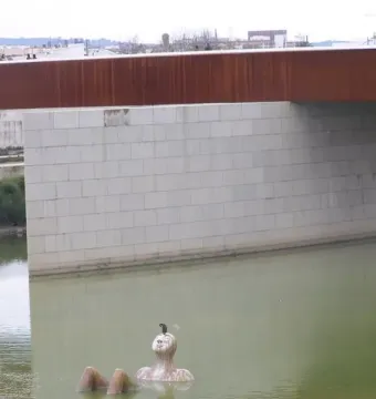 Hombre Río statue partly submerged in water beside a bridge pier, with a bird perched on its head.