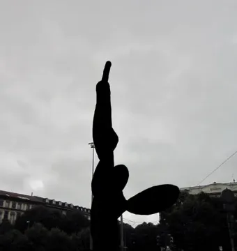 Silhouetted Needle, Thread and Knot sculpture rising against a cloudy sky in a public square.