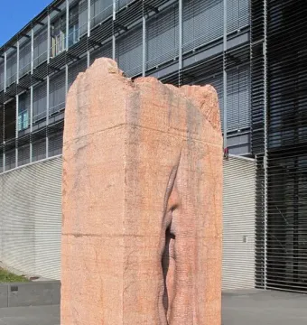 Pi-Chacán, a reddish-orange stone sculpture with a deep vertical crevice, standing on dark gravel beside a modern building.