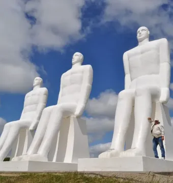 The Man by the Sea sculpture with four massive seated white figures against a blue sky, with a person beside the front statue.