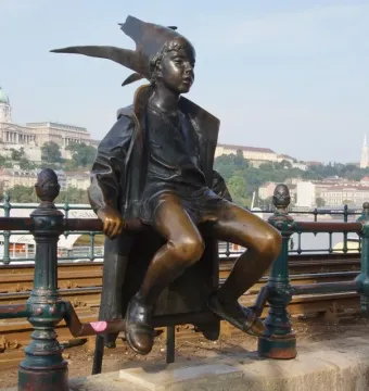 Little Princess bronze statue of a child sitting on a green railing with a city skyline and river in the background.