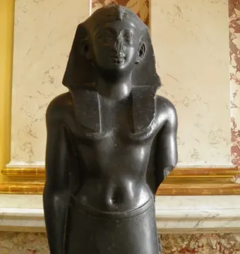 Upper body of a Ptolemaic king statue in dark stone, wearing an Egyptian headcloth and kilt with broken arms.