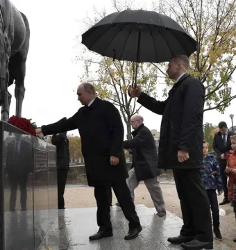 A man places flowers at the base of the Monument to the Russian Expeditionary Force while another person holds an umbrella nearby.