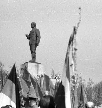 Stalin Monument with a uniformed figure gesturing from a tall pedestal above a crowd holding flags.