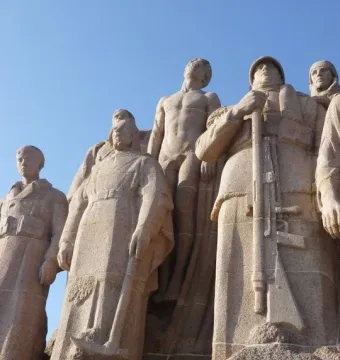 Stone figures from The Ghosts stand closely together, one holding a rifle and another a pickaxe, seen from below against the sky.