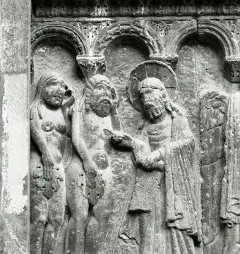 Stone relief of a haloed bearded figure pointing toward two nude figures beneath arched niches.