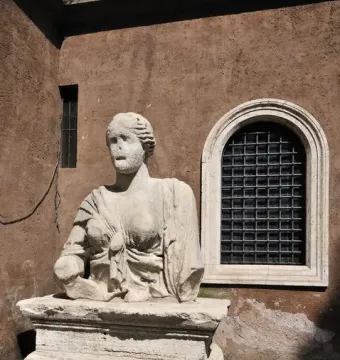 Madama Lucrezia stone bust of a woman in classical drapery beside a reddish-brown wall and arched window.