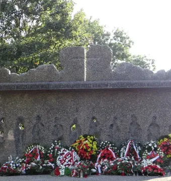 Monument to the Victims of the Wola Massacre with a rough memorial wall, carved figures, circular openings, and wreaths at the base.