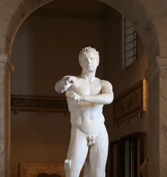 Apoxyomenos, a white marble nude male statue, shown under a museum archway scraping his left forearm.