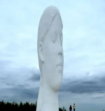 Dream sculpture, a tall white head and neck with closed eyes, standing on a circular base under a cloudy sky.
