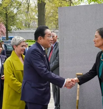 A man and a woman shake hands in front of the Japanese American Memorial, with several people standing nearby and blooming trees in the background.