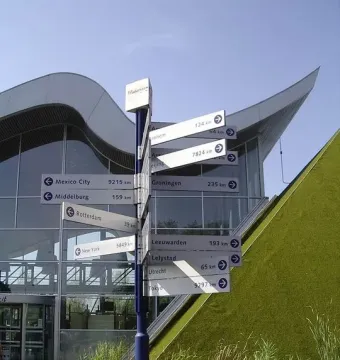 Madurodam signpost pointing to cities worldwide beside a modern building with a wavy roof and a pond in front.