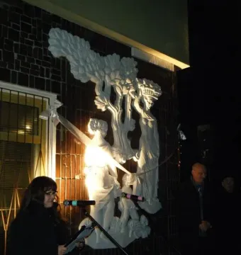 Plon relief sculpture mounted on a dark brick wall, with a woman speaking at microphones in front of it.