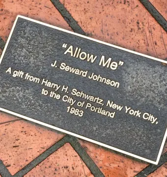 Dark metal plaque for Allow Me mounted on a brick pavement.