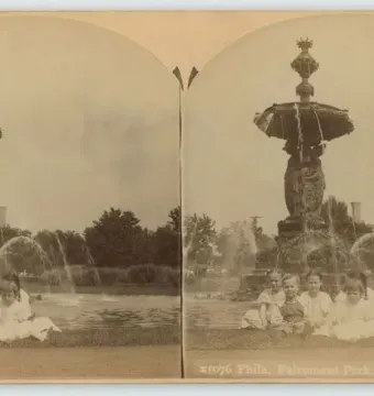 Children seated on the grass in front of the Bartholdi Fountain with water flowing into a surrounding pool.