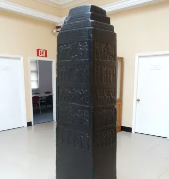 Black Obelisk with stepped top and carved relief bands on a wooden base in a museum interior.