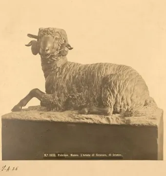 A bronze ram sculpture with curled horns rests on a dark pedestal with a small label near the base.