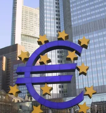 Blue euro sculpture surrounded by twelve yellow stars in front of modern buildings.