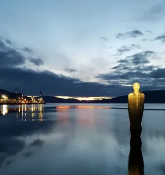Havmannen sculpture standing in calm water at twilight, with its reflection visible and an illuminated industrial area in the background.