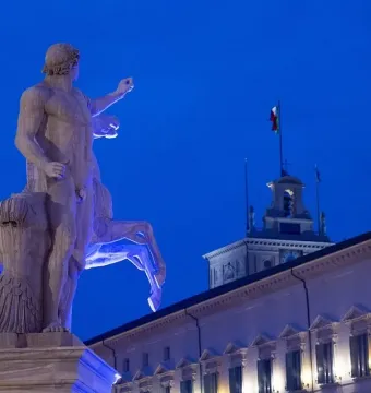 Horse Tamers sculpture lit in blue beside a horse, with an ornate building and clock tower in the background.