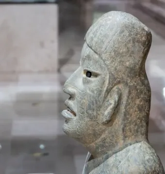 Side profile of the Las Limas Figurine showing a green-grey stone head with an open mouth, visible tongue, and hollow eye socket.