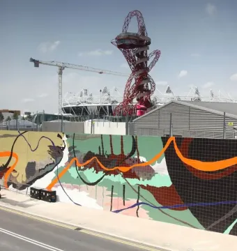 Orbit Tower rises in the background above a brightly tiled mural, with nearby buildings, a stadium, and a crane under a clear sky.