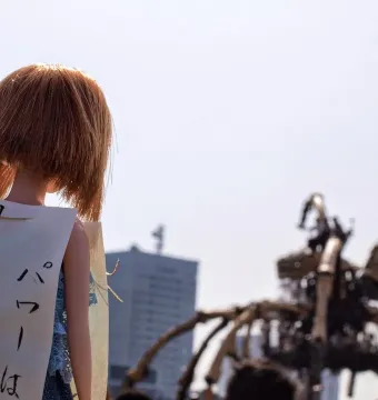 Doll with reddish-brown hair and a Japanese sign in front of La Princesse, with the large mechanical spider blurred in the background.