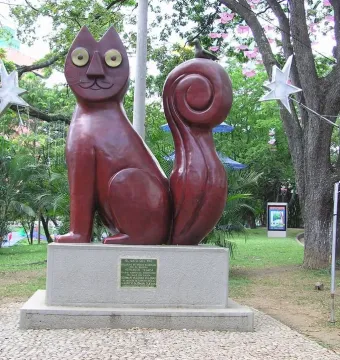 El Gato del Río cat sculpture on a stone pedestal in a park, with a small bird perched on its curled tail.