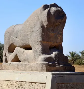 Lion of Babylon stone sculpture with a human figure beneath it, set against a blue sky and palm trees.