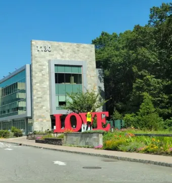 Red LOVE sculpture with a tilted O stands in front of a modern stone and glass building, surrounded by flowerbeds and greenery.