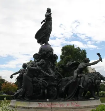 The Triumph of the Republic bronze monument with a central female figure standing on a globe, surrounded by dynamic figures and lions, on a stone base marked with red graffiti.