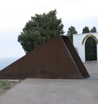 Walter Benjamin memorial sculpture in Portbou beside the cemetery archway, with the sea and trees in the background.