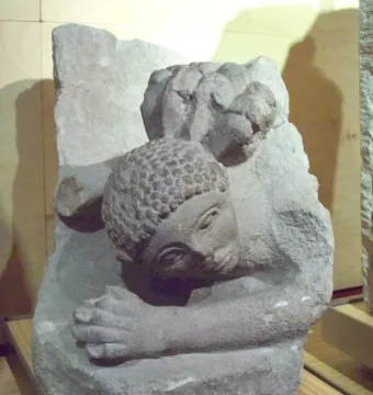 Stone relief of the Acrobat from Osuna, showing a figure with curled hair and clasped hands emerging from a rough block of stone.