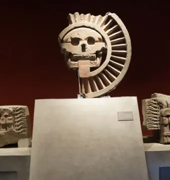 Disk of Mictlāntēcutli displayed with a carved stone skull and flanking stone heads in a museum exhibit.